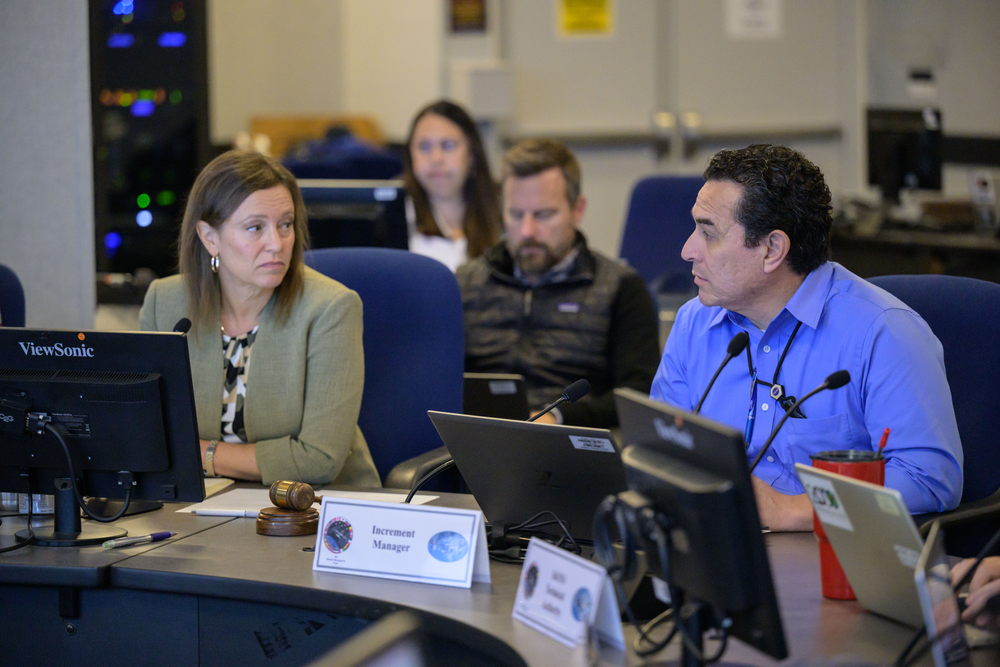 Two people sitting at a desk with computers in front of them and other people in the background. The person on the left is wearing a beige blouse and the person on the right is wearing a blue button down.